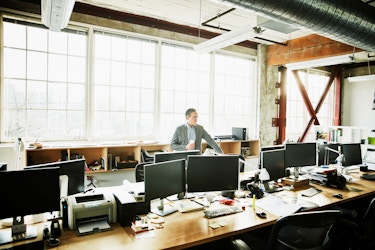  A business owner stands in a sunlit modern office that is empty of workers. He is surveying the workspace and weighing the merits of a business decision. 