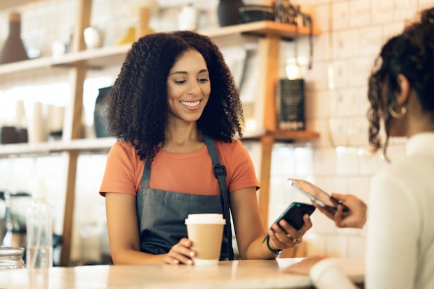 A coffee shop customer holds up her smartphone to a point-of-sale card reader to pay for her beverage.