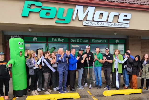 The front of a brick-and-mortar PayMore store during a store opening. The signage above the store is the PayMore logo -- "Pay" in green and "More" in white. About two dozen store employees stand in front of the store, applauding. One store employee is dressed as a neon green inflatable tube man. The words "We Buy Sell & Trade Electronics" stretch across the top of the glass doors leading into the store.