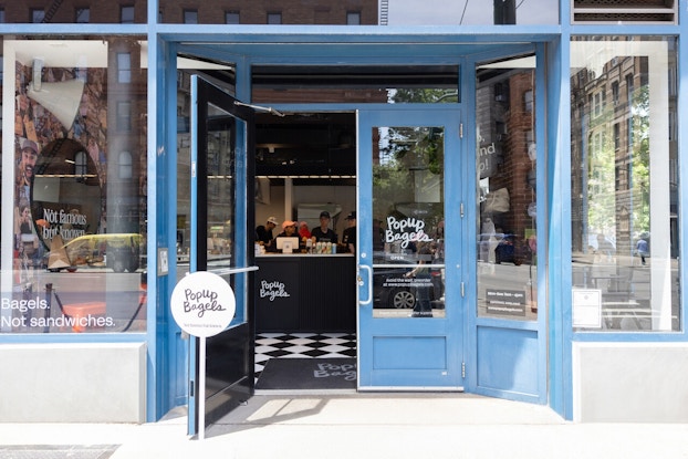The exterior of a PopUp Bagels location. The double-doors and windows of the shop are outlined in blue, and the name of the shop is written on the glass of the doors in a playful cursive script. One door stands open, revealing the shop's black-and-white checkerboard floors and a checkout counter with staff standing behind it.