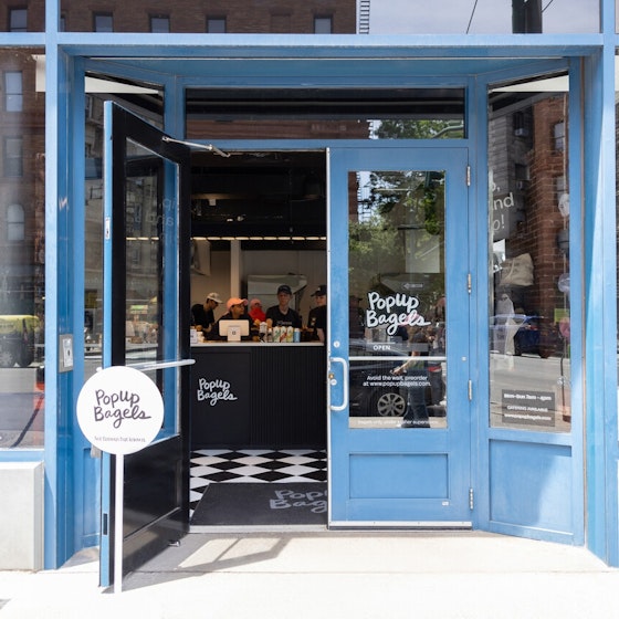 The exterior of a PopUp Bagels location. The double-doors and windows of the shop are outlined in blue, and the name of the shop is written on the glass of the doors in a playful cursive script. One door stands open, revealing the shop's black-and-white checkerboard floors and a checkout counter with staff standing behind it.