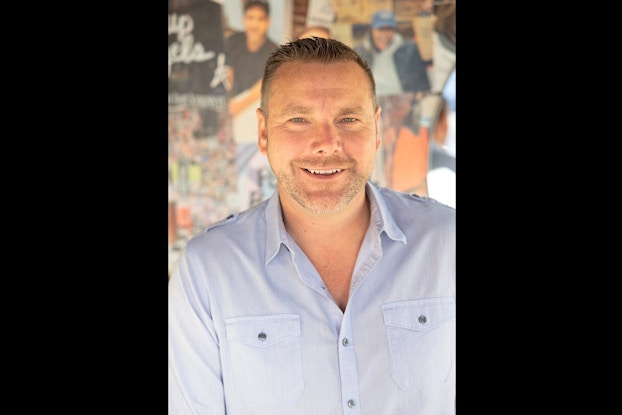 A headshot of PopUp Bagels CEO Tory Bartlett, smiling for the camera. Bartlett is a middle-aged white man with short dark hair and a light beard. He wears a pale blue button-up shirt.