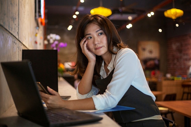A cafe owner sits at a counter in her shop. She has a laptop open in front of her and is holding her smartphone. Her cafe is empty and she looks worried.
