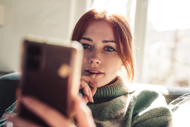 A woman with red hair is seated on a couch. The image features a close-up of her face. She is holding a smartphone in front of her and is looking at something on the phone screen.