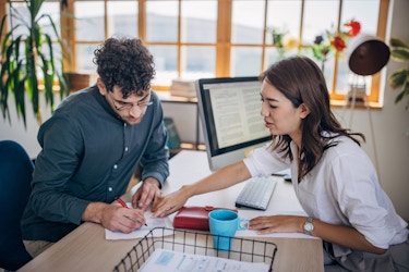 Two people are shown in a bank lender's office. The man, a small business owner, is signing loan documents while the woman, a loan officer, is pointing out which areas of the loan agreement he needs to acknowledge with a signature.