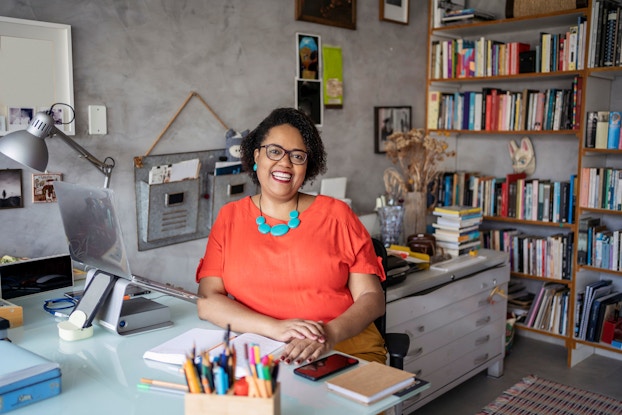 A mature Black woman is pictured seated at her desk in a cozy office. She is smiling at the viewer.
