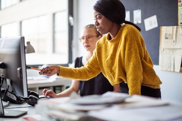 Two business professionals look at a computer monitor and discuss an issue. A woman points at the computer monitor and talks with a female colleague.