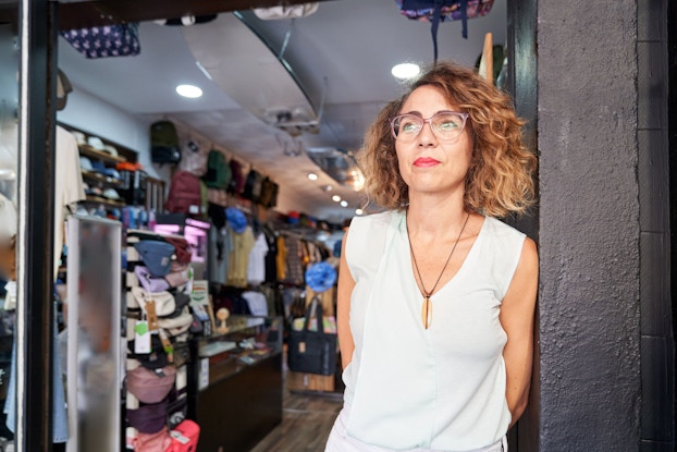 A woman retail shop owner stands at the entrance to her shop. She is waiting to greet customer to her store.