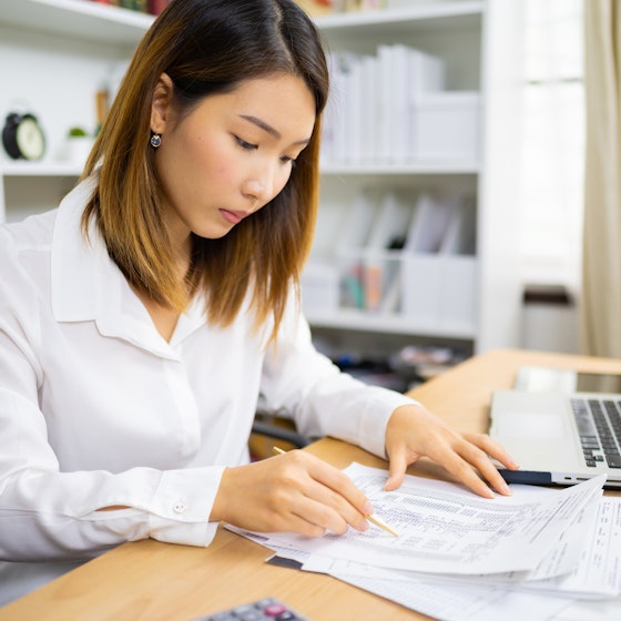 A woman solopreneur is seated at a desk in her home office. She is reviewing a spreadsheet in front of an open laptop computer. She is tallying deductions for her income taxes.