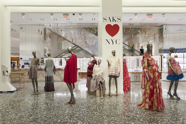  Nine mannequins model bright summer dresses. Several of the mannequins are clustered around a column bearing the message "SAKS HEARTS NYC."