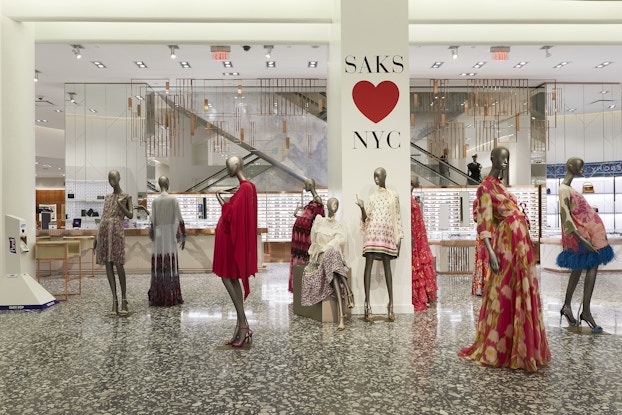 Nine mannequins model bright summer dresses. Several of the mannequins are clustered around a column bearing the message "SAKS HEARTS NYC."