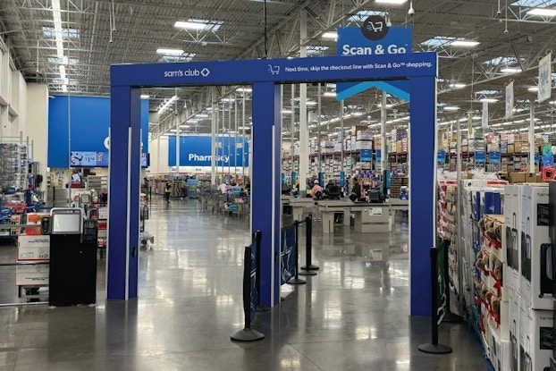 The entry of a Sam's Club, with two blue Scan & Go overhead sensors in the shape of oversized doorways through which shoppers must pass to enter and exit. The left sensor has the Sam's Club logo on top, while the right sensor has the words "Next time, skip the checkout line with Scan & Go shopping" written across the top. In the background is the enormous Sam's Club warehouse, with a blue Scan & Go sign hanging from the ceiling near the checkout area.