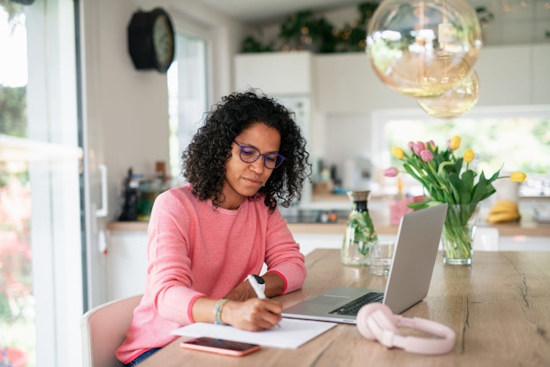  A young woman seated in her sunny kitchen with her laptop in front of her takes notes on a pad of paper.