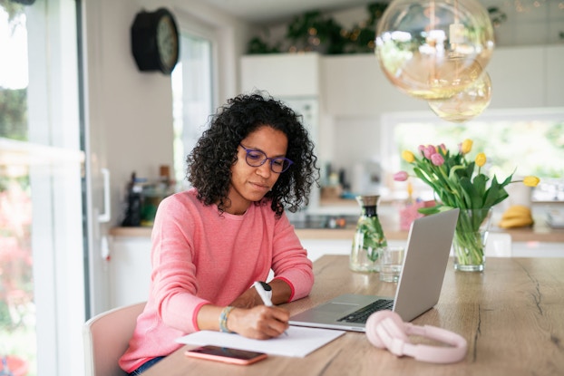A young woman seated in her sunny kitchen with her laptop in front of her takes notes on a pad of paper.