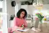 A young woman seated in her sunny kitchen with her laptop in front of her takes notes on a pad of paper.