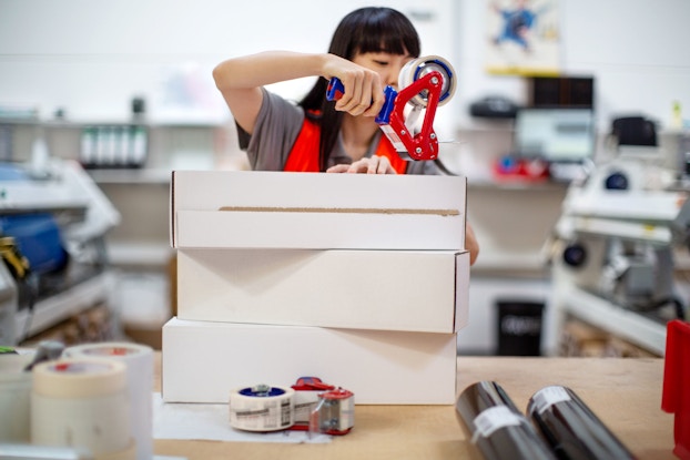  A female worker preparing boxes for shipment. She is sealing a box with shipping tape.