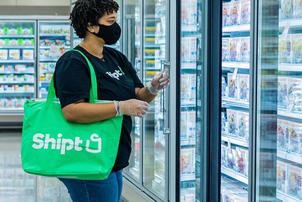  A Shipt employee wearing a mask looks through the frozen food section of a partnering grocery store for requested food items, which will be placed in the large green bag hanging from the shopper's shoulder.