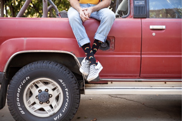  A man sits on the side of the bed of a red truck. His torso and face are not pictured, bringing focus to his cuffed, light denim pants and exposed are his decorative black crew socks.