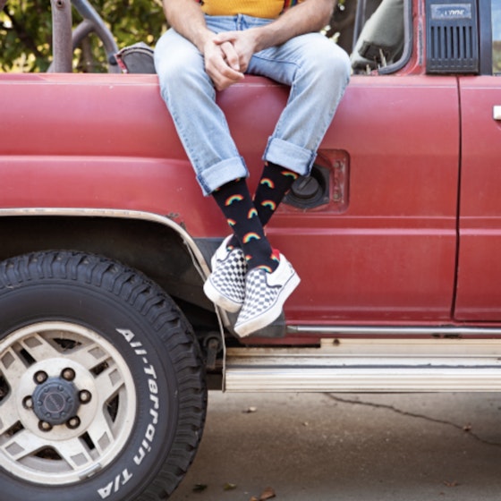  A man sits on the side of the bed of a red truck. His torso and face are not pictured, bringing focus to his cuffed, light denim pants and exposed are his decorative black crew socks. 