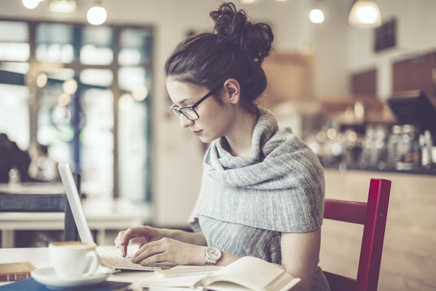  Young woman wearing glasses works on laptop in cafe. On the desk is a coffee cup and open book.