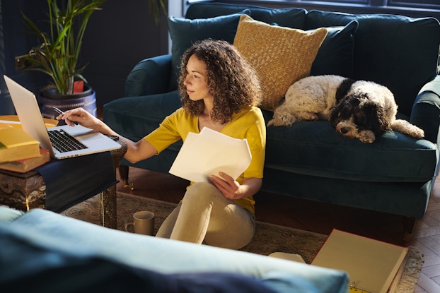 A woman sits on her living room floor with papers and a laptop to do work. Her dog lays on the couch near her.