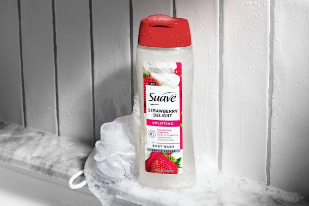 A bottle of Suave strawberry shower gel and a loofah sit on a marble shelf in a bath.