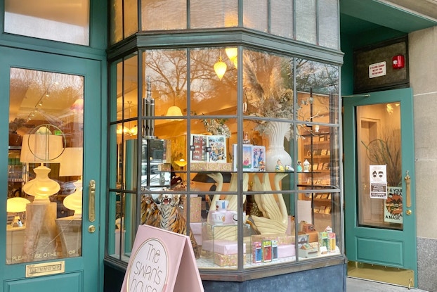 Portrait of a shop's exterior with two green doors and a bay window, the shop's sign sits out front on the sidewalk.