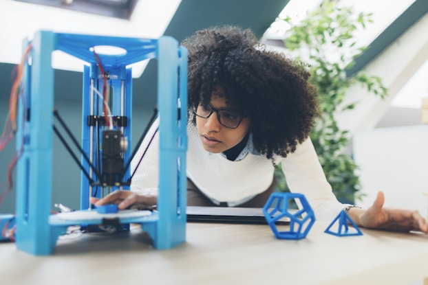  Person working in office, leaning over desk and 3D printer.