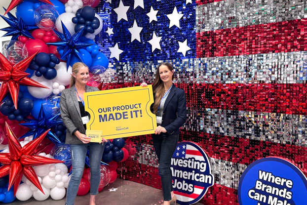 Jordan Catapano and Jocelyn Dunn, Co-founders of This Girl Walks into a Bar, with their golden ticket from Walmart's Open Call pitch competition. The two woman stand in front of a massive bedazzled American flag and red-white-and-blue balloons. They're holding a large sign shaped like a golden ticket reading "OUR PRODUCT MADE IT!" In the background are two other signs reading "Investing in American Jobs" and "Where Brand Can Make It Big."