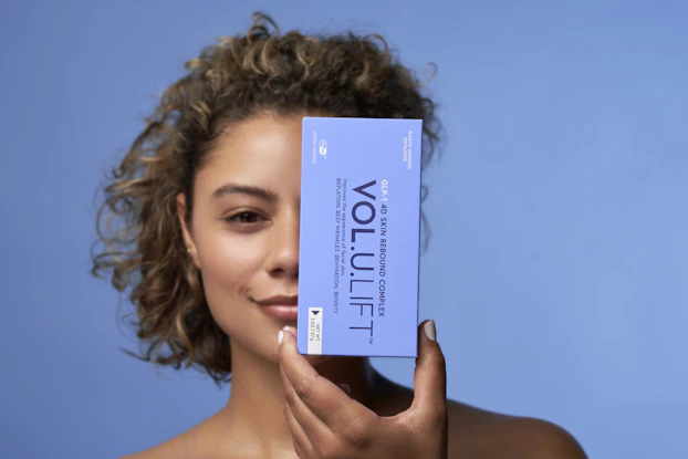 A woman holding up a rectangular blue package of Image Skincare's Vol.U.Lift, obscuring half of her face.