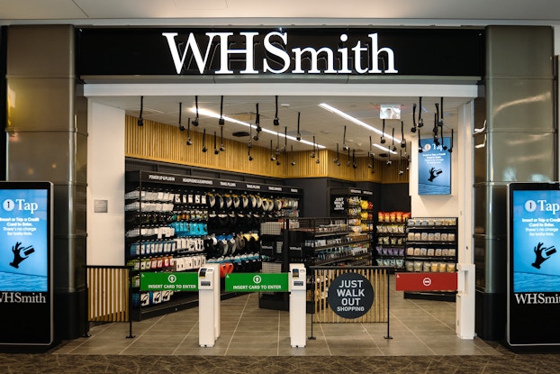 The entry of a WHSmith outlet at an airport. The entry is flanked by two LED screens showing how to use WHSmith's "Just Walk Out" automatic checkout system. The entry is partially blocked by grating, one portion of which has a round sign that reads "JUST WALK OUT Shopping." Three gates leads into the store itself; two of the gates are green and labeled "Insert Card to Enter," and third gate is red, to stop shoppers from entering through an exit. Inside, a couple dozen cameras and sensors hang from the store's ceiling to capture people's purchases. The store's walls and floor displays are filled with shelves of snacks, drinks, electronic chargers, headphones, and other travel necessities.