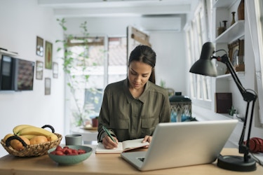 Young woman makes notes in a notebook from her computer while at a desk.