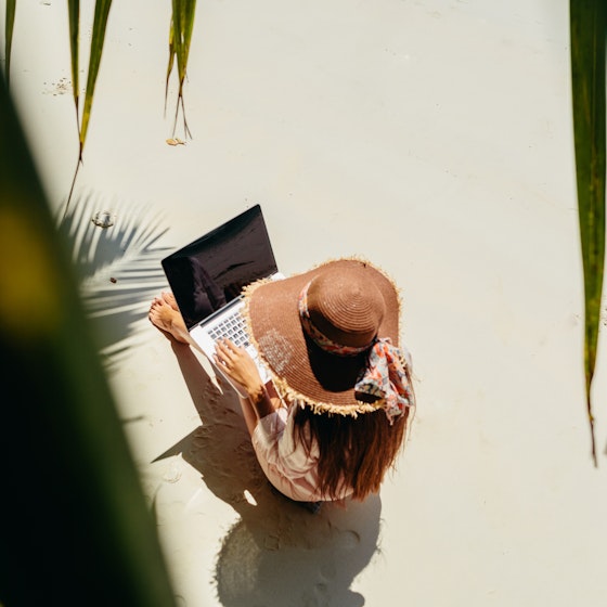 Top view of a woman working on her laptop while sitting in the sun on white sand.