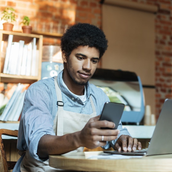 Man in apron sits at table and works on smartphone and laptop in interior of loft coffee shop.