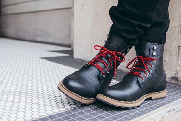 A person wearing black boots with tan soles and red laces stands on a tile floor