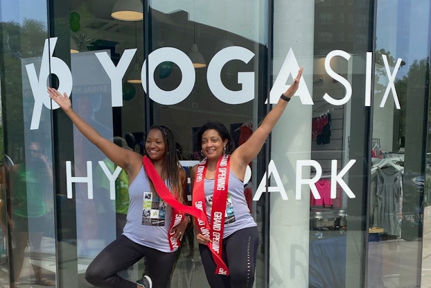 Two women standing in front of a yoga studio in a yoga pose, standing on one leg with one arm stretched over head.