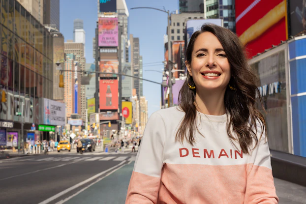 Julie Chapon, Co-founder and CEO of Yuka, smiling and standing in New York City's Time Square. Julie is a white woman with long wavy brown hair and she wears a white-and-pink pullover shirt and gold hoop earrings.
