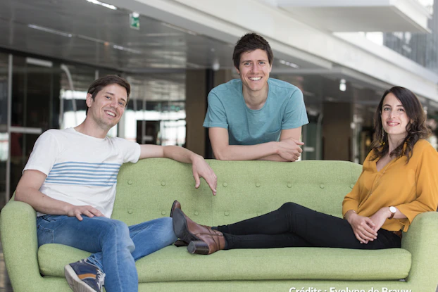 The three Co-founders of Yuka smiling as they sit and lean on a light green couch. From left to right, they are Benoit Martin sitting with his legs crossed and one arm across the back of the couch; Francois Martin leaning over the back of the couch; and Julie Chapon sitting with her legs stretched out on the couch.