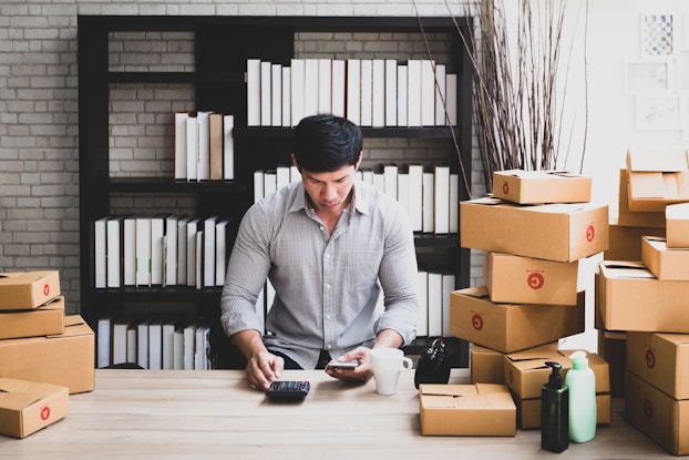 A man stands behind a counter piled with cardboard boxes. The boxes are taped closed and many are marked with red circles containing either the letter A or the letter C. The man is looking down the smartphone held in his hand and a calculator on the desk.