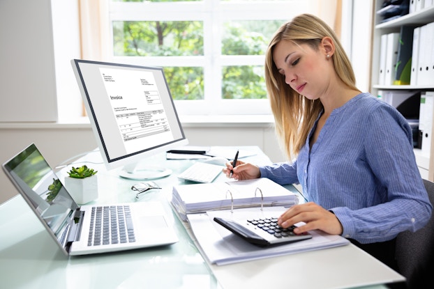 A woman consults a calculator while writing something in a binder full of papers. Both a laptop and a computer monitor sit on the desk near her, with the monitor displaying an invoice..