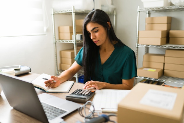 A woman sits at a desk in a storage room, writing in a notebook with one hand while typing on a calculator with the other. The woman has long dark hair and is wearing a dark teal blouse. Behind her are metal shelving units filled with cardboard boxes.