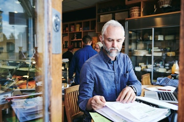  A deli business owner sits at a table in his deli. Before him he has his laptop computer and a binder filled with invoices and receipts. He is thumbing through the invoices and reviewing data as he prepares to update his business's books. 