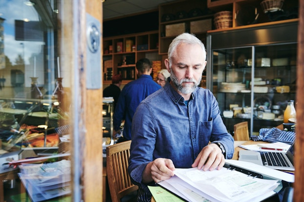  A deli business owner sits at a table in his deli. Before him he has his laptop computer and a binder filled with invoices and receipts. He is thumbing through the invoices and reviewing data as he prepares to update his business's books.