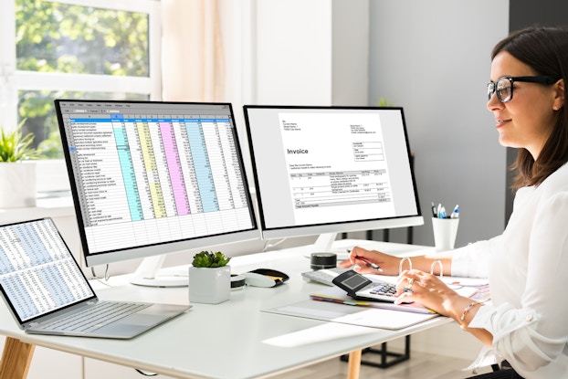 A woman consults a laptop and two computer monitors, which show spreadsheets and invoices. The woman types something on a calculator.