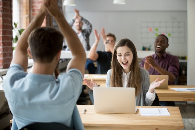Several people in an office celebrate. The camera is focused on a woman in business casual wear looking at something on a laptop with an open-mouthed smile on her face and her arms spread in surprise and celebration. Behind and in front of her, her colleagues are seated at wood-topped tables and applauding.