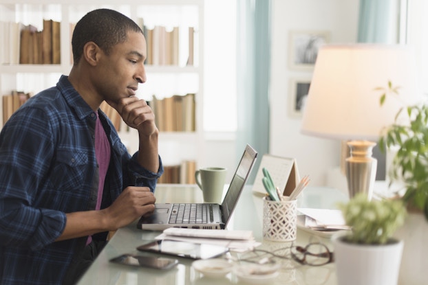  A man sits at a white desk and looks at the screen of an open laptop. Scattered elsewhere on the desk are a couple of potted plants, a cup of pencils and scissors, a tablet and smartphone, and a few stacks of papers. The man is wearing a dark blue plaid shirt over a purple T-shirt, and he has one hand held to his chin in thought.