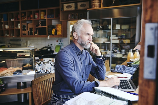  An older man sits at a table in the front room of a deli. The man has white hair and a graying beard and he wears a dark blue button-up shirt. He has one hand on the keyboard of a laptop and the other held to his chin in deep thought. Next to the laptop is an open binder filled with pages of text. Behind the man are glass-fronted display cases filled with slices of deli meat and a glass-fronted refrigerator filled with wheels of cheese.