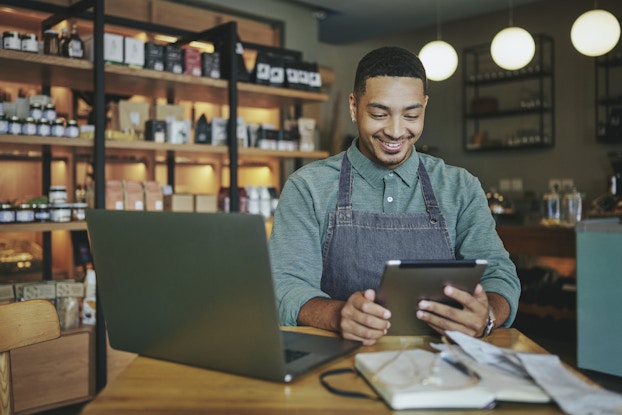  A man in a denim apron sits at a wooden table and looks down at the electronic tablet in his hands with a smile. The man has dark hair shaved close to his head and some light facial hair. On the table in front of him are an open laptop and an open notebook with a pair of glasses sitting on top of it. In the background is a gourmet food shop. One wall is lined with shelving units filled with jars, boxes, and bags of prepared foods. Globular light fixtures hang from the ceiling.