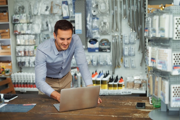 A man stands behind a worn wooden checkout desk and leans forward to type on a small laptop. The man has short brown hair and wears a pale blue button-up shirt and khakis. He is smiling at whatever he sees on the laptop's screen. Behind him is a wall of hardware store merchandise, including coiled and uncoiled metal tubes, various sized bottles of glue, and small items individually wrapped in plastic. A spinning stand holding similar items sits on top of the checkout counter.