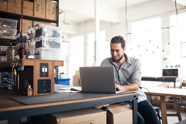 A man sits at a counter in a large workshop space. He types something on a laptop and looks down at the laptop screen with a smile. The man is young and bearded, and he is wearing a blue-gray button-up shirt. The counter next from him has a shelf holding several clear plastic boxes, and stacks of cardboard boxes sit under the counter and on metal shelves beside the counter. In the background are rows of long tables and a white wall with several large windows letting in light.